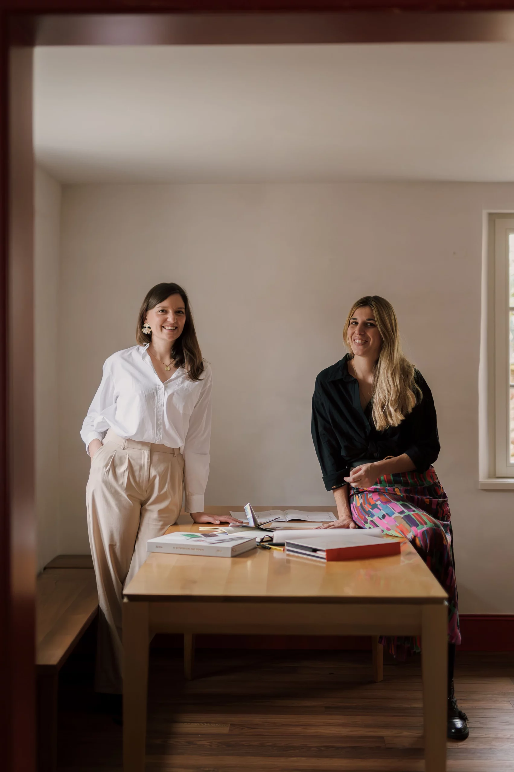 Deux femmes souriantes travaillant ensemble dans un bureau lumineux, entourées de livres et de documents.